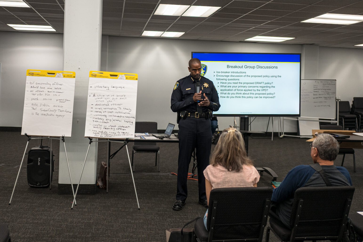 Uniformed police chief speaks to a small group during a community meeting, standing between two flip-charts and a screen labeled “Breakout Group Discussions.”