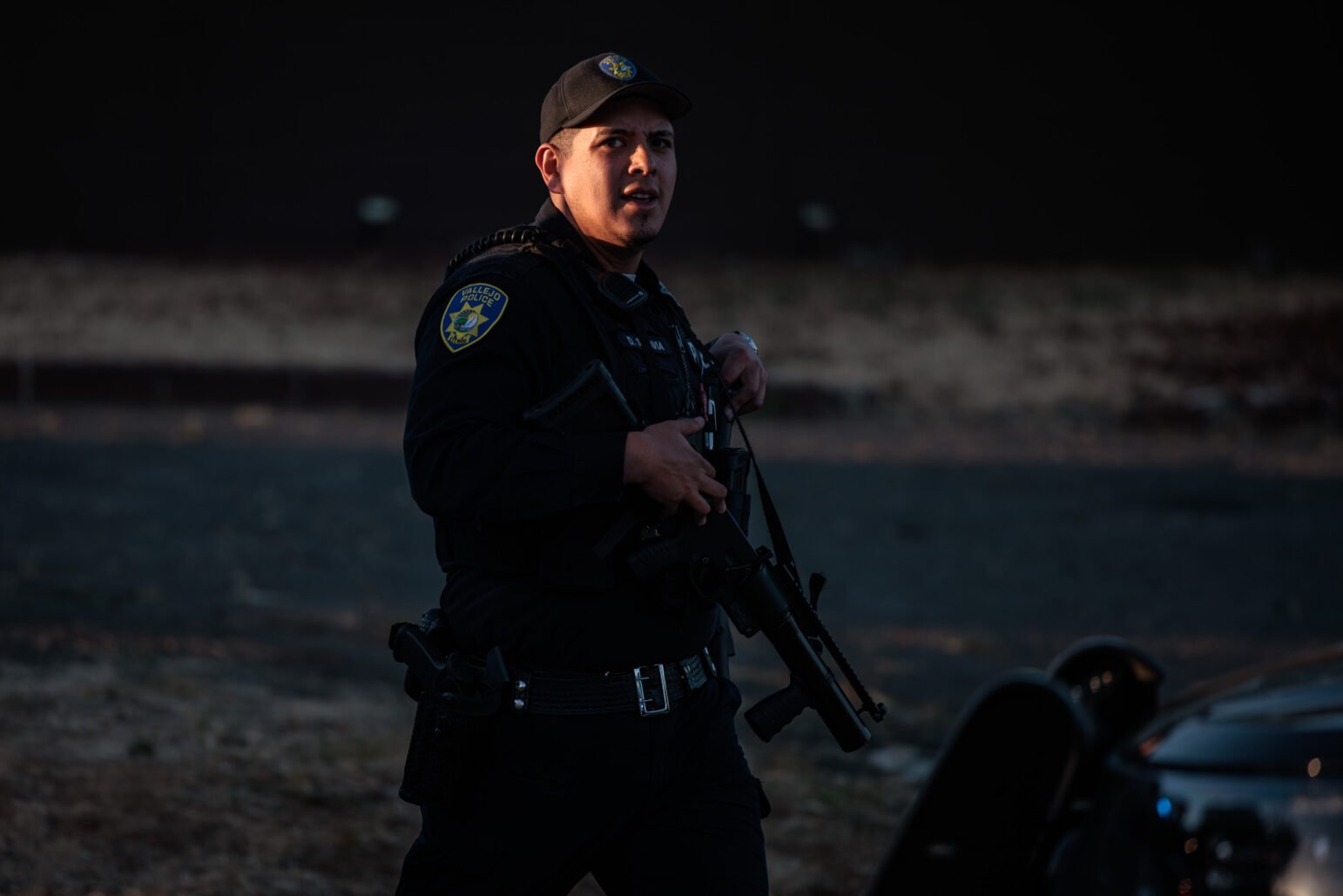 A uniformed officer wearing a cap walks while carrying a less-lethal weapon near a police vehicle, lit by low evening light.
