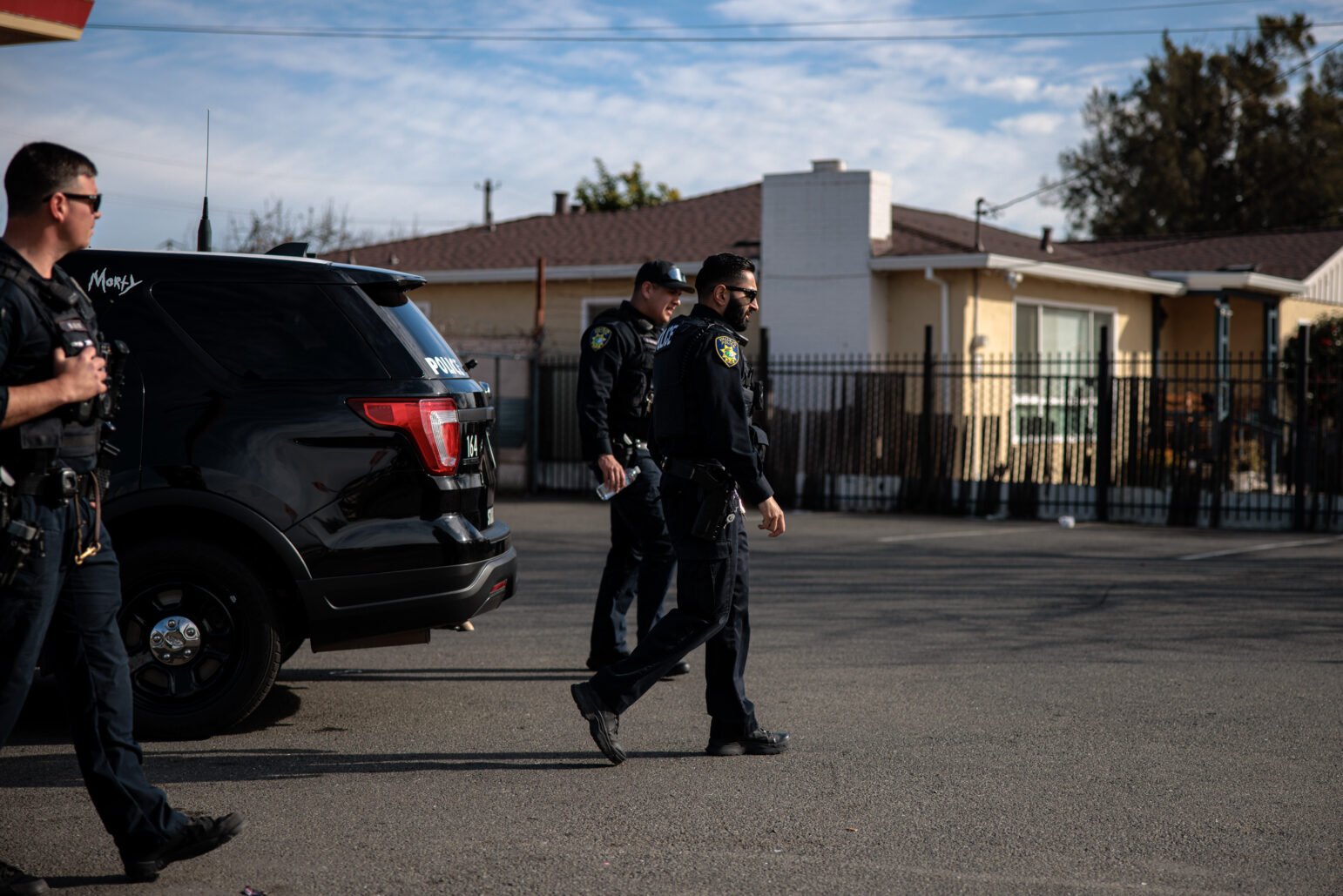Three police officers walk past a parked SUV in a parking lot, with single-story houses and a black metal fence in the background.