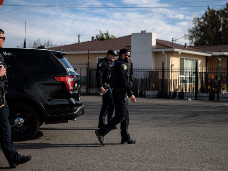 Three police officers walk past a parked SUV in a parking lot, with single-story houses and a black metal fence in the background.