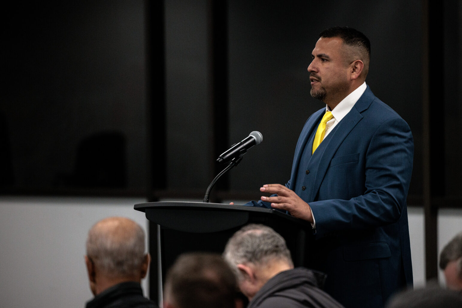 Man in a blue suit and yellow tie addresses the room from a lectern with a microphone, audience heads visible in the foreground.