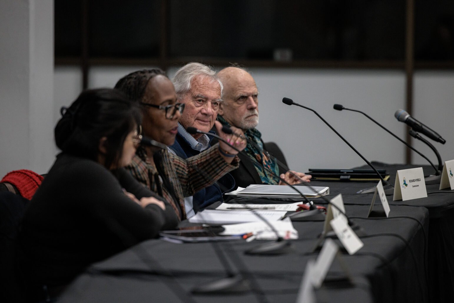 Four commissioners sit at a long table with microphones; one woman gestures while the others listen during a public meeting.