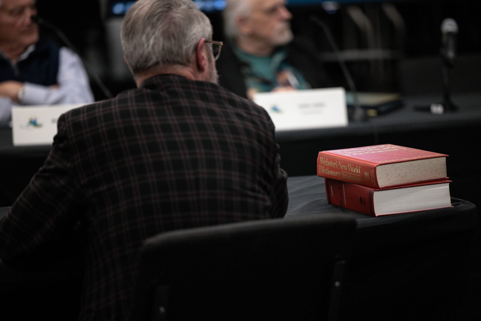 Two red dictionaries stacked on a table in the foreground, with blurred panelists and name placards in the background.