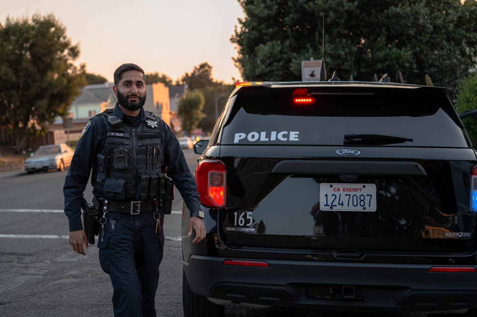 Officer in dark uniform and tactical vest stands beside a marked police SUV with rear lights in a residential street.