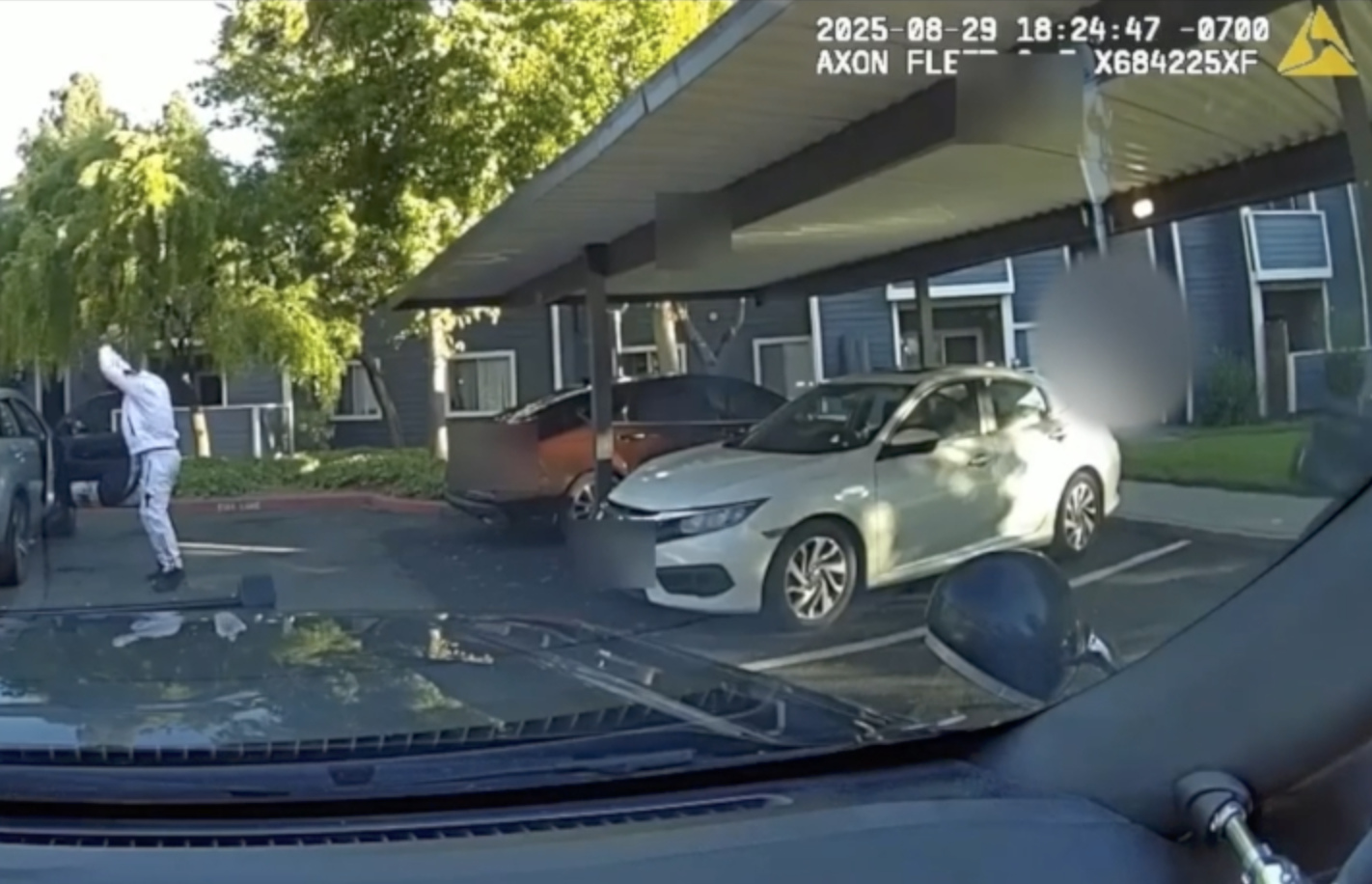 Police dashcam view of an apartment-complex parking lot; a person in a light tracksuit stands beside an open car door with his hands raised. Parked cars sit under a carport, a white sedan centered; parts of the image are blurred for privacy, a timestamp (2025-08-29 18:24:47 -0700) overlays the top right, and the patrol car’s dashboard and side mirror are visible.