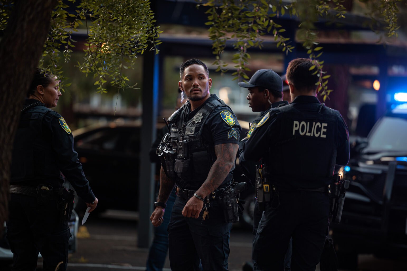 A group of police officers in tactical gear stand together outdoors near a police vehicle with flashing lights, one officer looking alertly to the side while others engage in conversation.