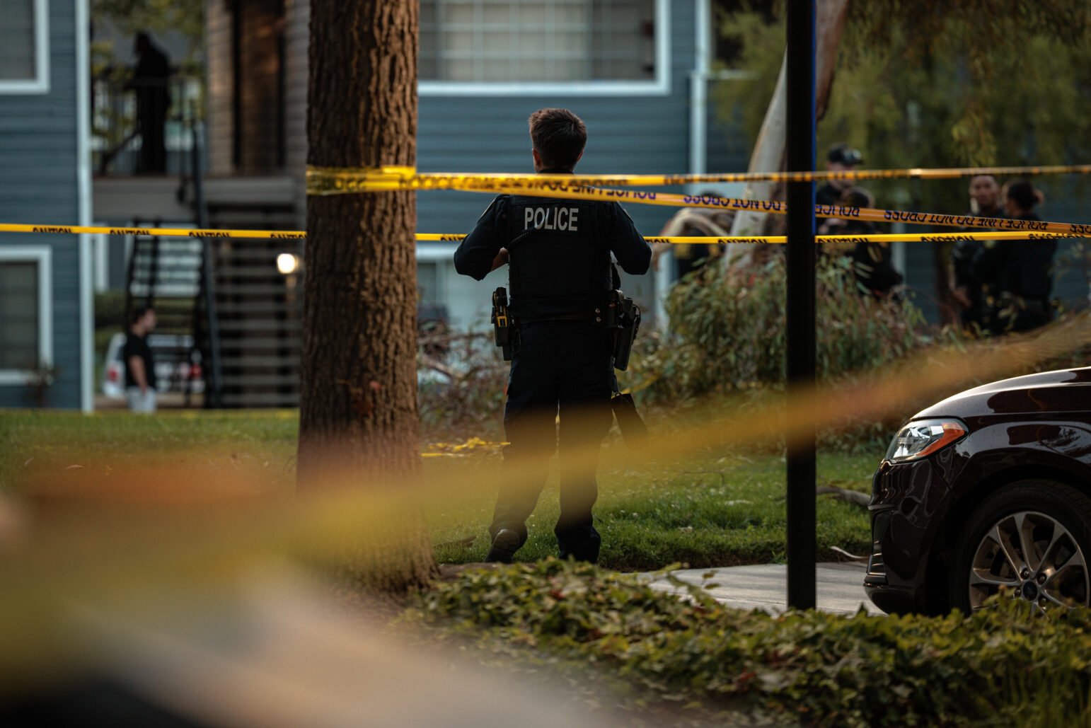 Police officer in uniform stands facing yellow caution tape at an outdoor scene in a residential area, with other people and officers visible in the background near apartment buildings and parked cars.