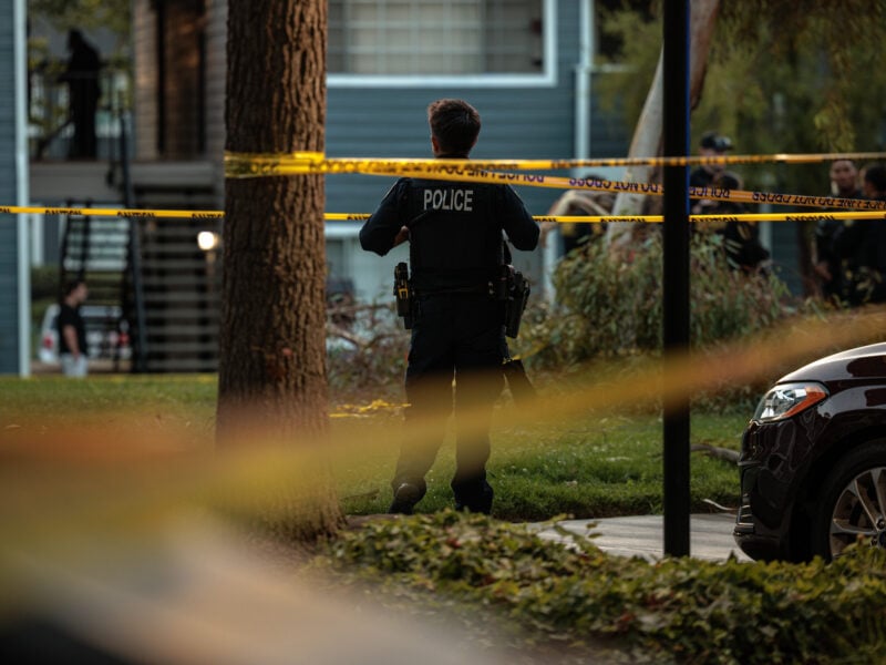 Police officer in uniform stands facing yellow caution tape at an outdoor scene in a residential area, with other people and officers visible in the background near apartment buildings and parked cars.
