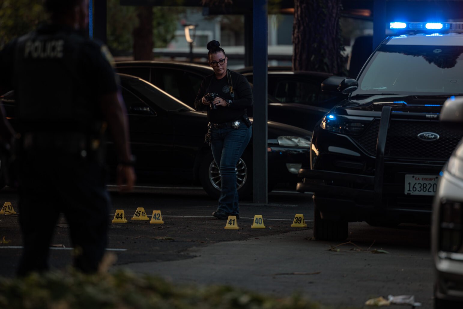 Police at a nighttime crime scene; yellow evidence markers numbered in the 30s and 40s line the pavement near a patrol SUV with blue lights.