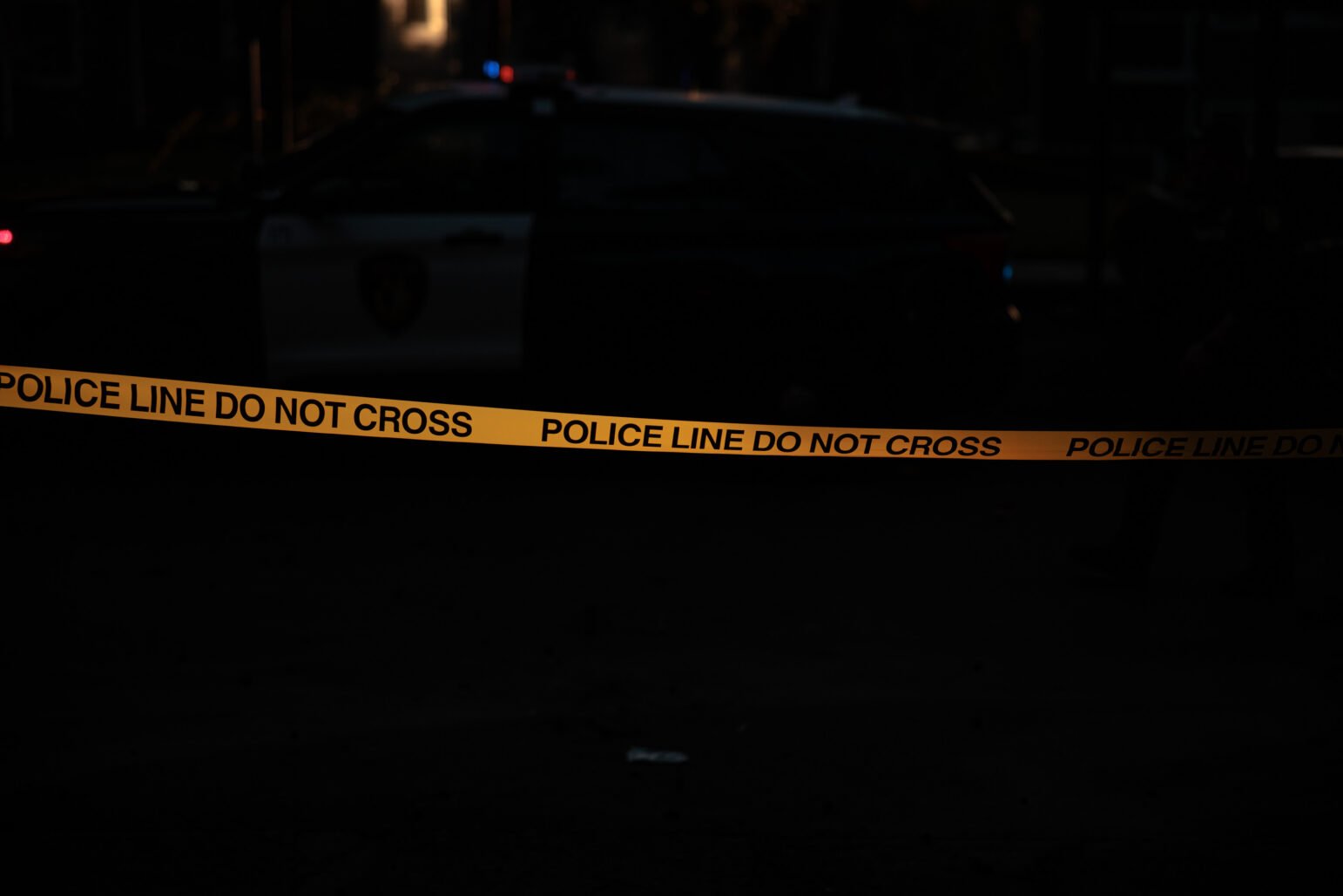 Yellow police tape reading “POLICE LINE DO NOT CROSS” stretches across a dark scene with a police vehicle faintly visible in the background.