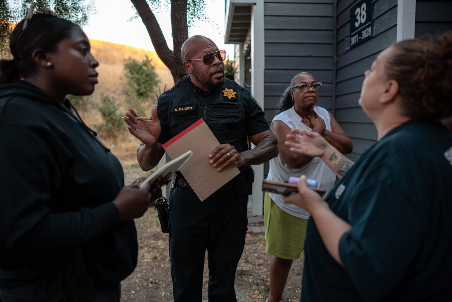 Outdoors, a uniformed officer wearing sunglasses holds a clipboard and talks with three women, one a detective in plainclothes, beside an apartment building.