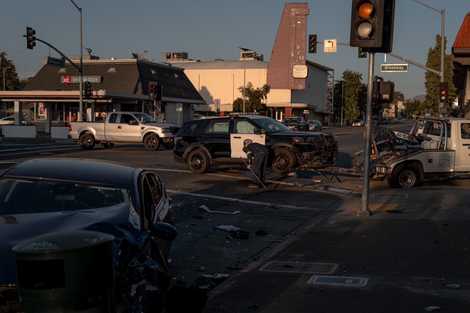 Aftermath of a crash at an urban intersection—black-and-white police SUV with heavy front-end damage near a tow truck while a worker in safety gear sweeps debris; a crumpled black car sits in the foreground with traffic lights and Broadway/Tennessee St signs and storefronts behind.