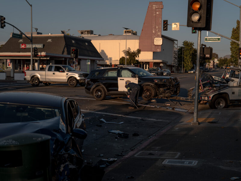 Aftermath of a crash at an urban intersection—black-and-white police SUV with heavy front-end damage near a tow truck while a worker in safety gear sweeps debris; a crumpled black car sits in the foreground with traffic lights and Broadway/Tennessee St signs and storefronts behind.