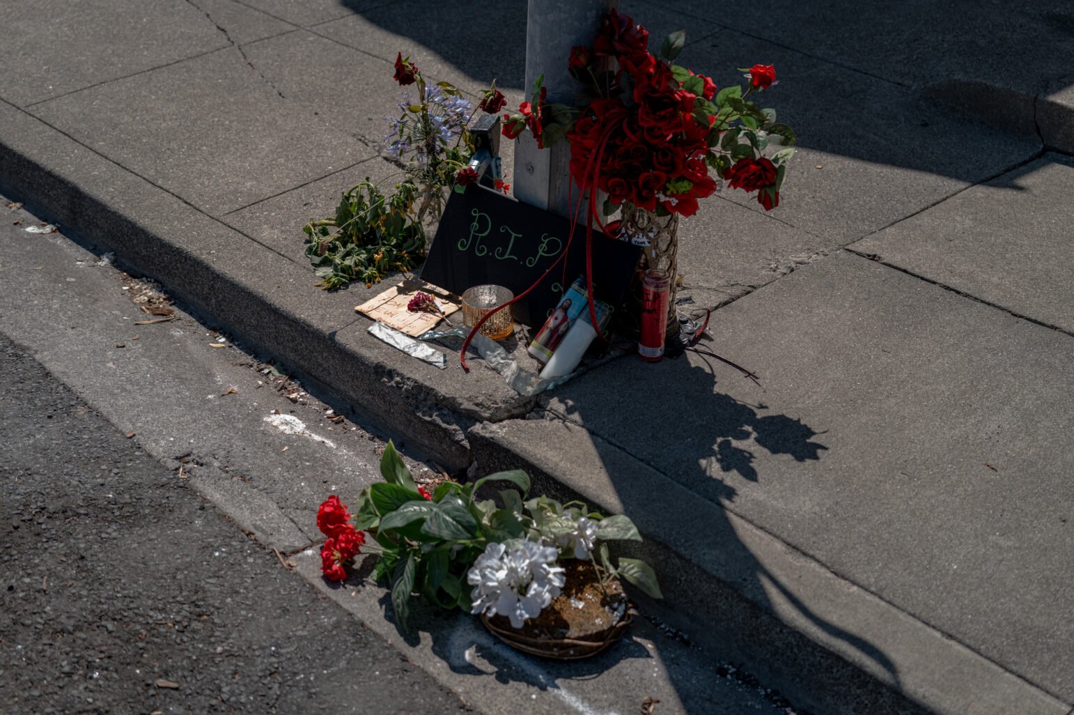 Small roadside memorial at the base of a street pole on a concrete corner—black board reading R.I.P., red roses with ribbon, prayer candles, wilted flowers, and small offerings scattered along the curb with sharp afternoon shadows.