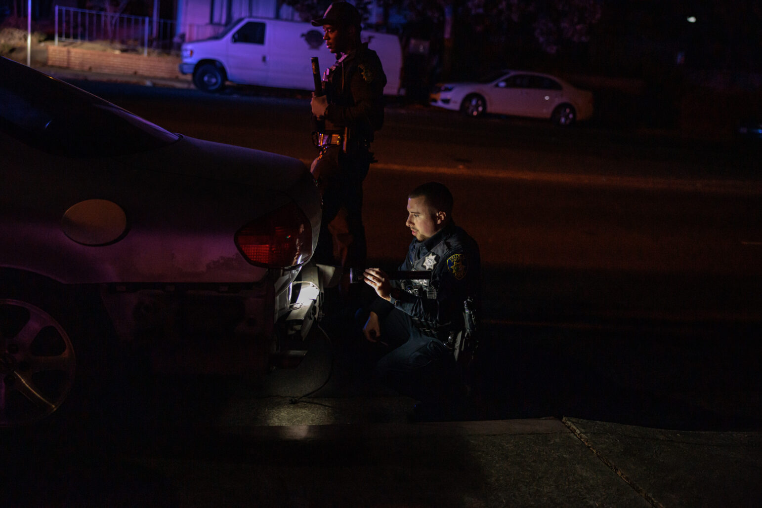 At night, a kneeling police officer shines a flashlight on the damaged rear bumper of a car while another officer stands watch beside him.