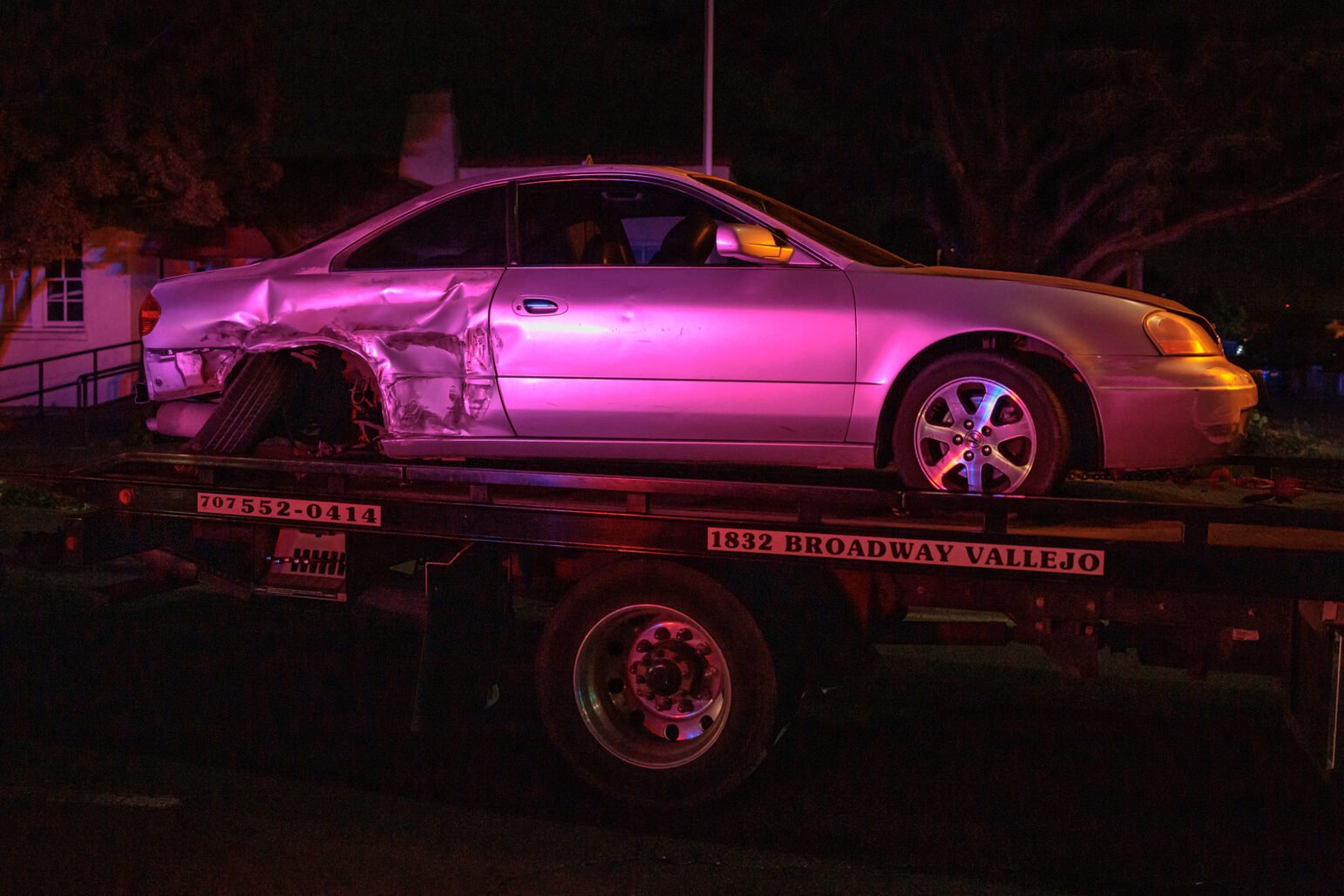 A silver coupe with heavy rear-side damage rests on a flatbed tow truck under flashing red and blue lights at night.