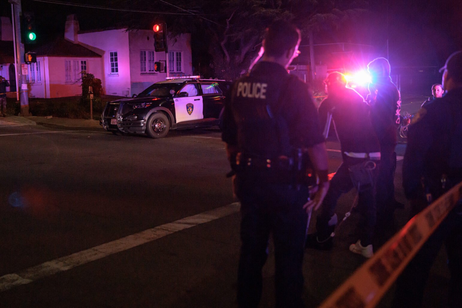 Nighttime intersection with police tape in the foreground, officers and several people near flashing lights, and a patrol SUV blocking the road.