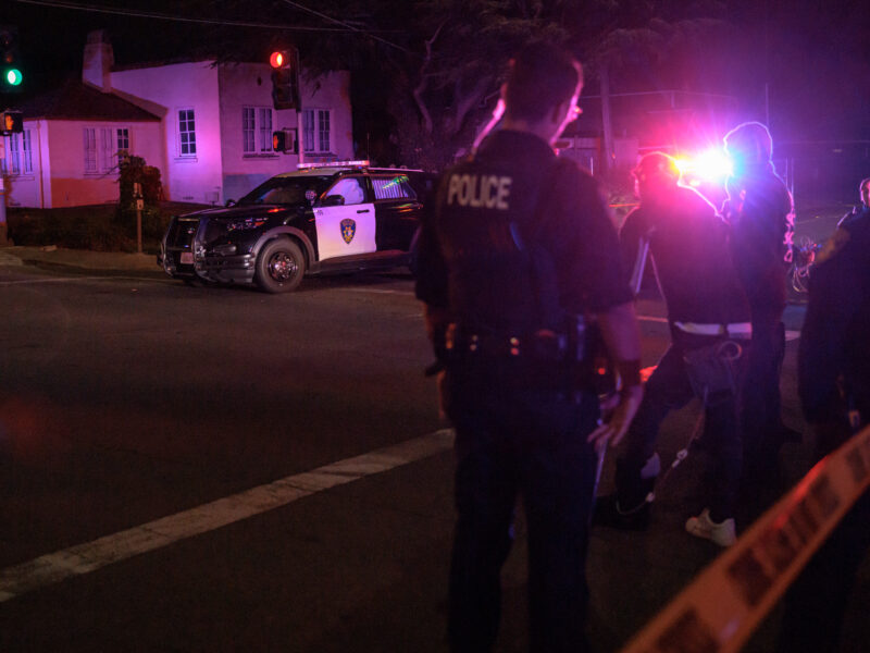 Nighttime intersection with police tape in the foreground, officers and several people near flashing lights, and a patrol SUV blocking the road.