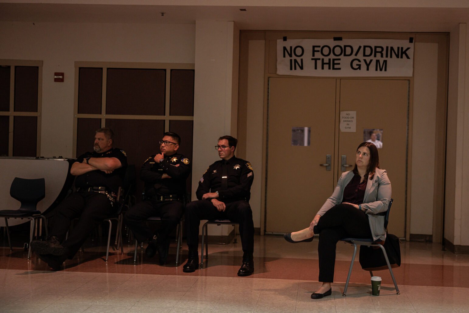 Three uniformed police officers and one woman in business attire sit in chairs inside a gymnasium, with a sign above the doors behind them that reads “NO FOOD/DRINK IN THE GYM.”