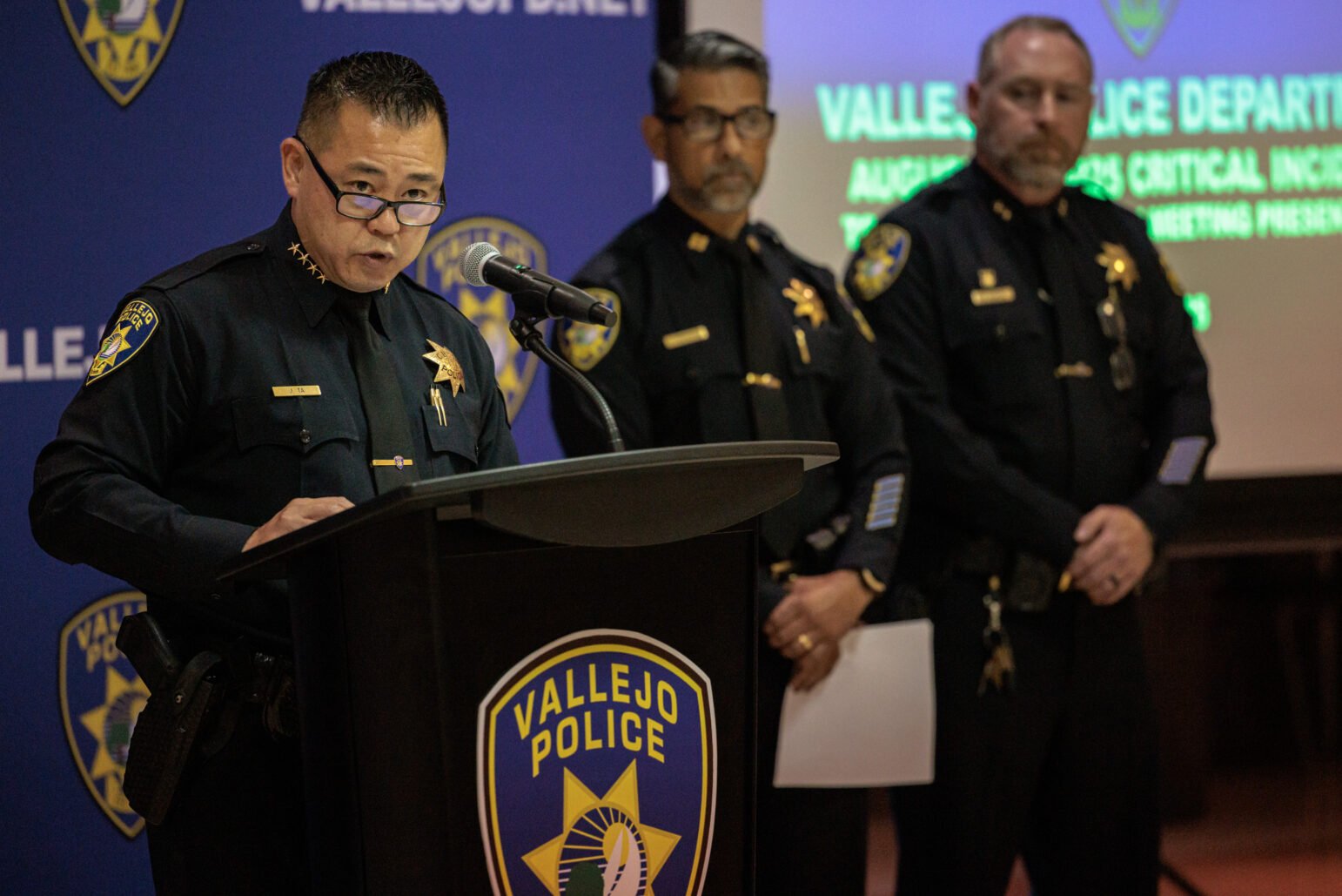 A uniformed police chief wearing glasses speaks at a Vallejo Police lectern with a microphone, while two other senior officers stand behind near a blue department backdrop and a projected screen.