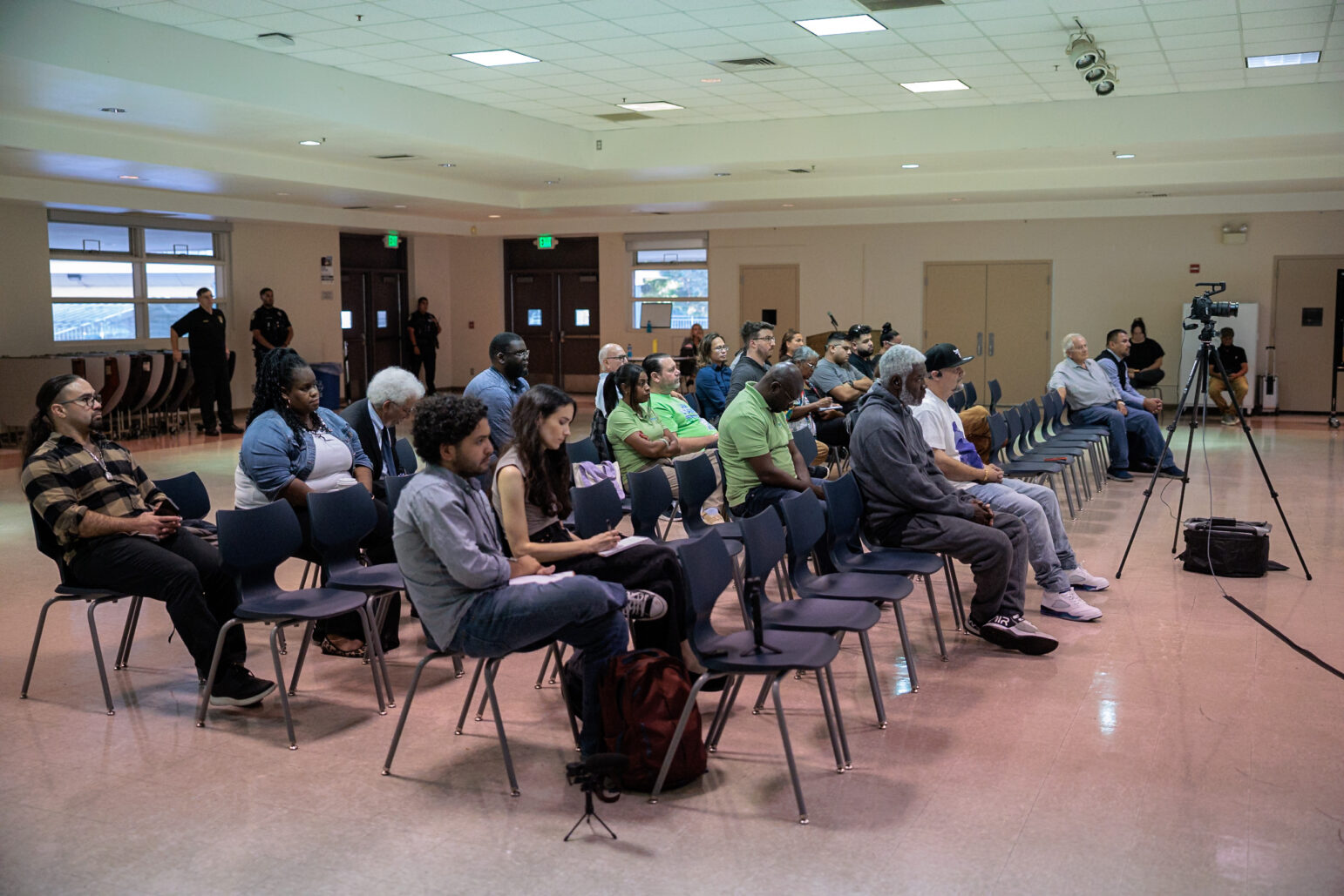 Audience members sit in rows in a multipurpose room for a community meeting, with a video camera on a tripod and officers standing along the back wall.
