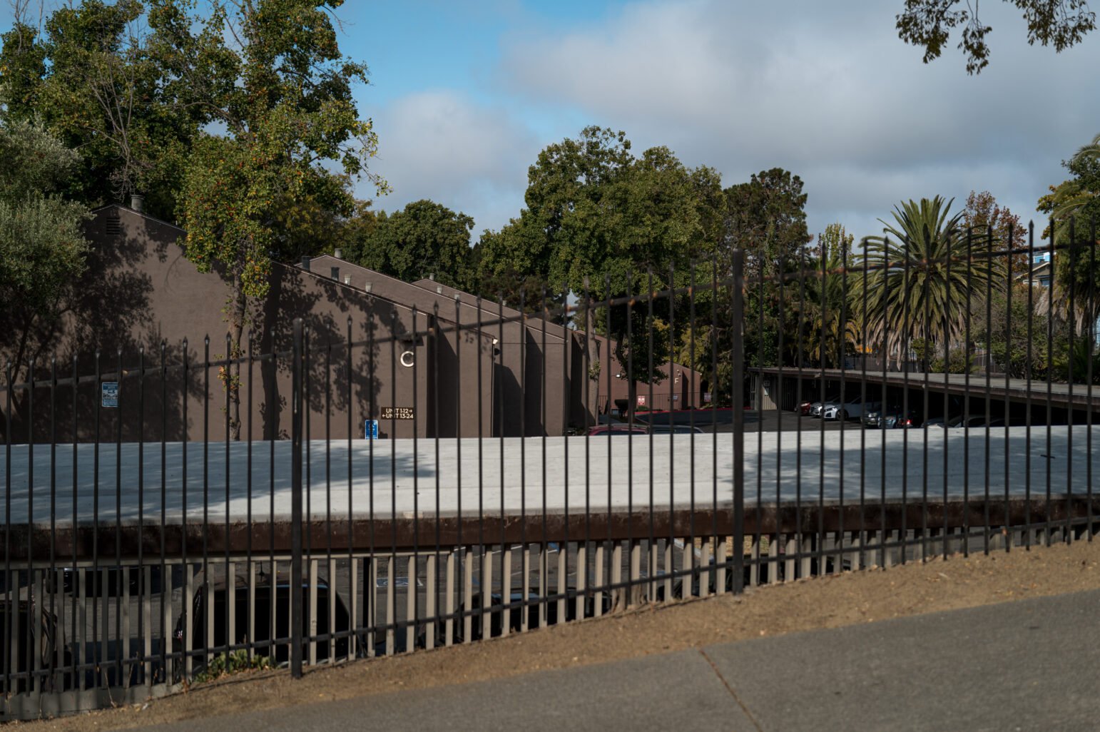 Black metal fence borders an empty rooftop parking deck with tan stucco buildings and trees, including a tall palm, under a partly cloudy blue sky.