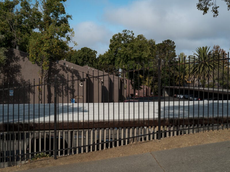 Black metal fence borders an empty rooftop parking deck with tan stucco buildings and trees, including a tall palm, under a partly cloudy blue sky.