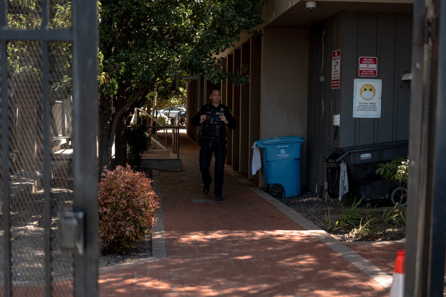 Police officer in uniform walks down a shaded brick walkway between buildings, framed by a gate; recycling and trash bins and “No Trespassing” signs line the wall, with shrubs and a traffic cone nearby.