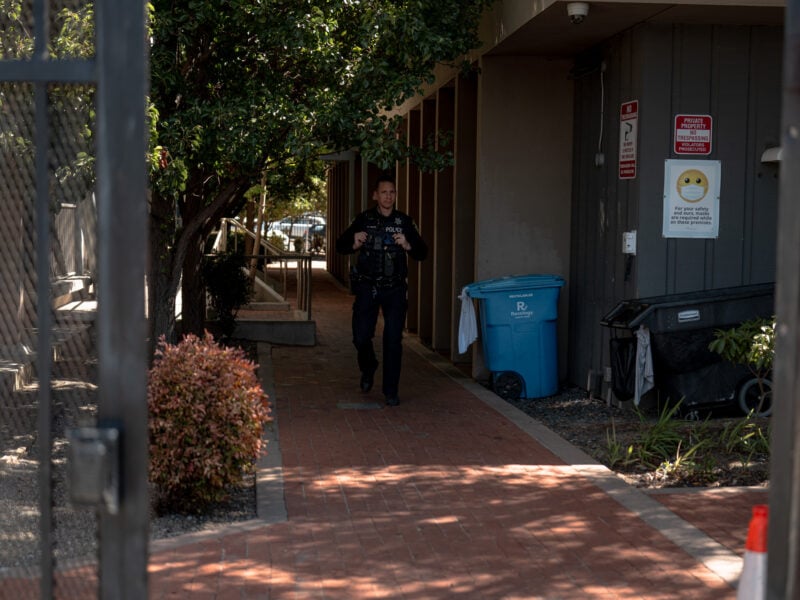 Police officer in uniform walks down a shaded brick walkway between buildings, framed by a gate; recycling and trash bins and “No Trespassing” signs line the wall, with shrubs and a traffic cone nearby.