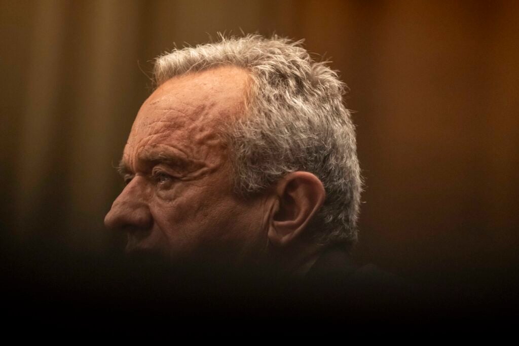 Close-up left-profile of an older man with short gray hair in warm, low light, his lined face in sharp focus while a dark blurred foreground obscures the lower frame.