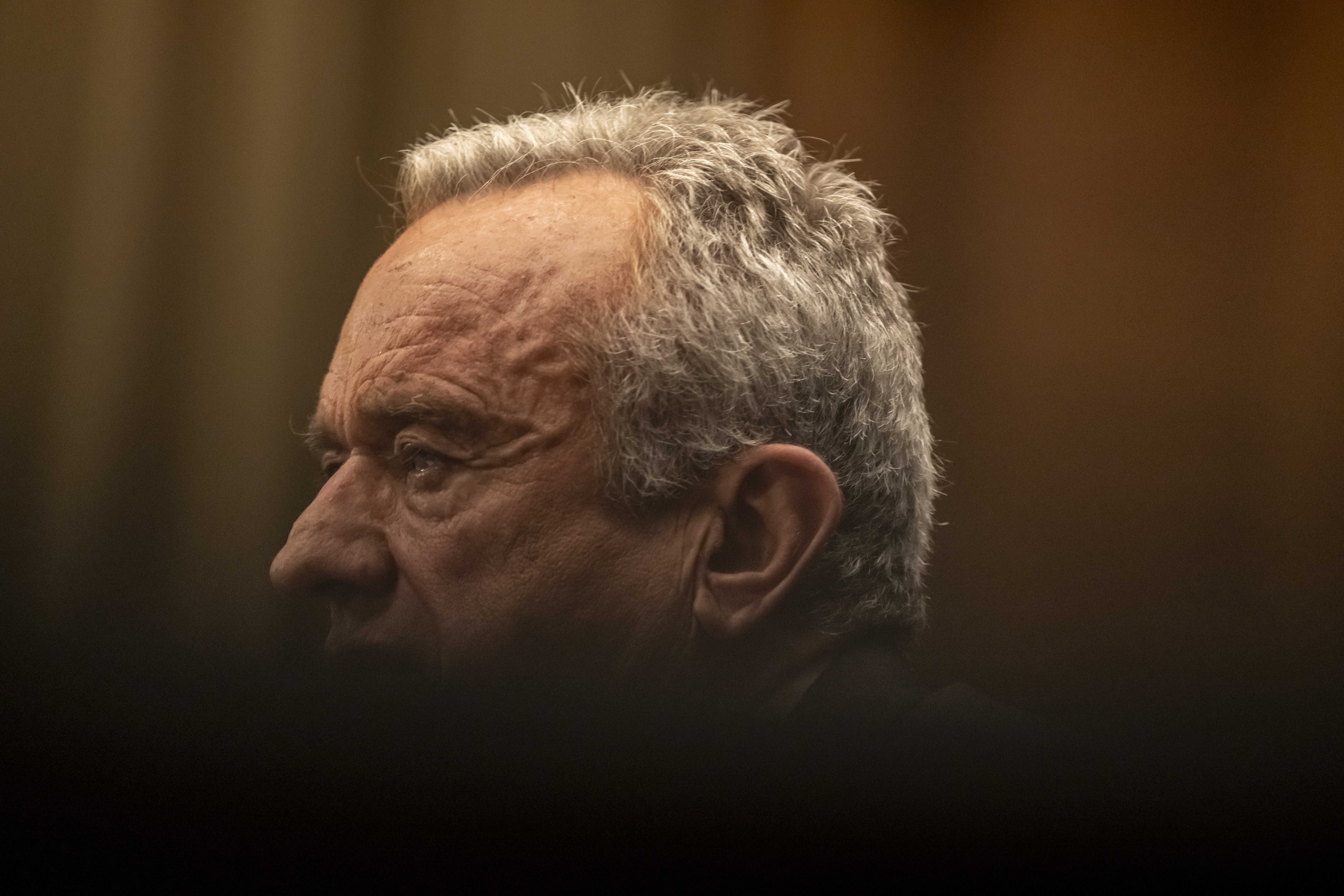 Close-up left-profile of an older man with short gray hair in warm, low light, his lined face in sharp focus while a dark blurred foreground obscures the lower frame.