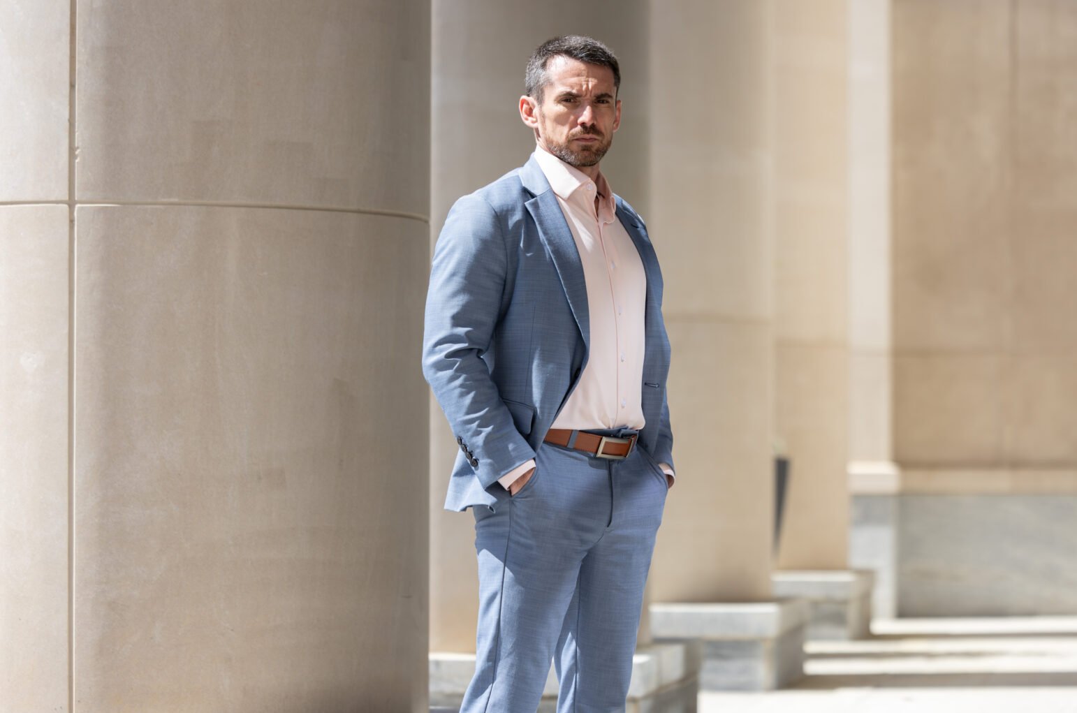 Man in a light blue suit and pale pink shirt stands with hands in pockets beside large stone columns, looking toward the camera in bright daylight.