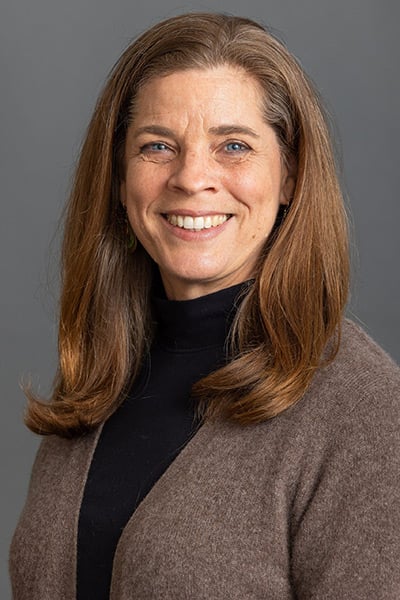 Headshot of a smiling woman with shoulder-length light brown hair, wearing a black turtleneck and brown cardigan against a gray background.