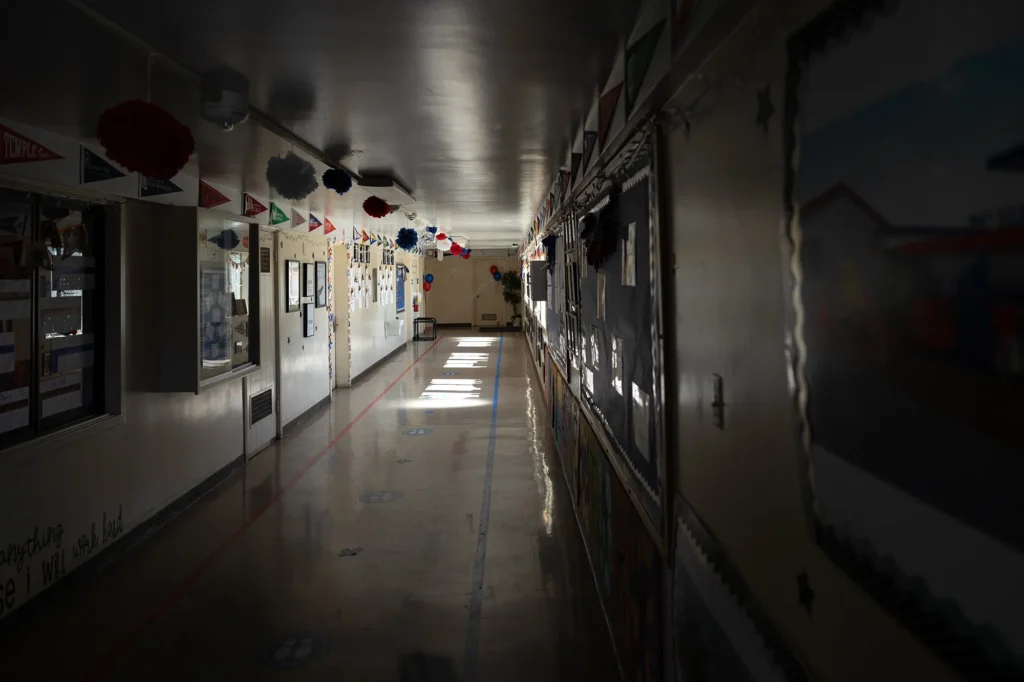 Empty, dimly lit school hallway with decorated bulletin boards and pennant banners; patches of sunlight on the floor lead toward a plant at the far end.