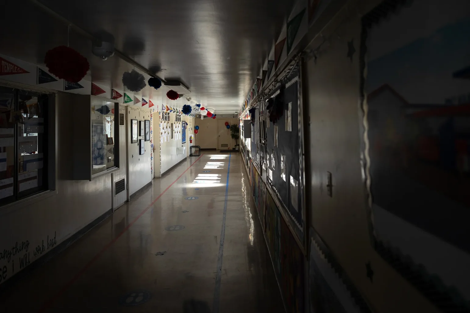 Empty, dimly lit school hallway with decorated bulletin boards and pennant banners; patches of sunlight on the floor lead toward a plant at the far end.