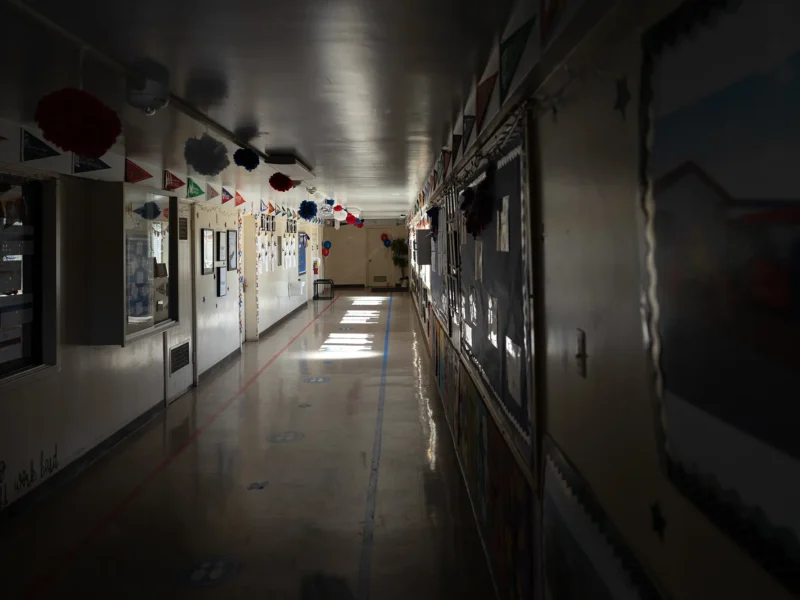 Empty, dimly lit school hallway with decorated bulletin boards and pennant banners; patches of sunlight on the floor lead toward a plant at the far end.