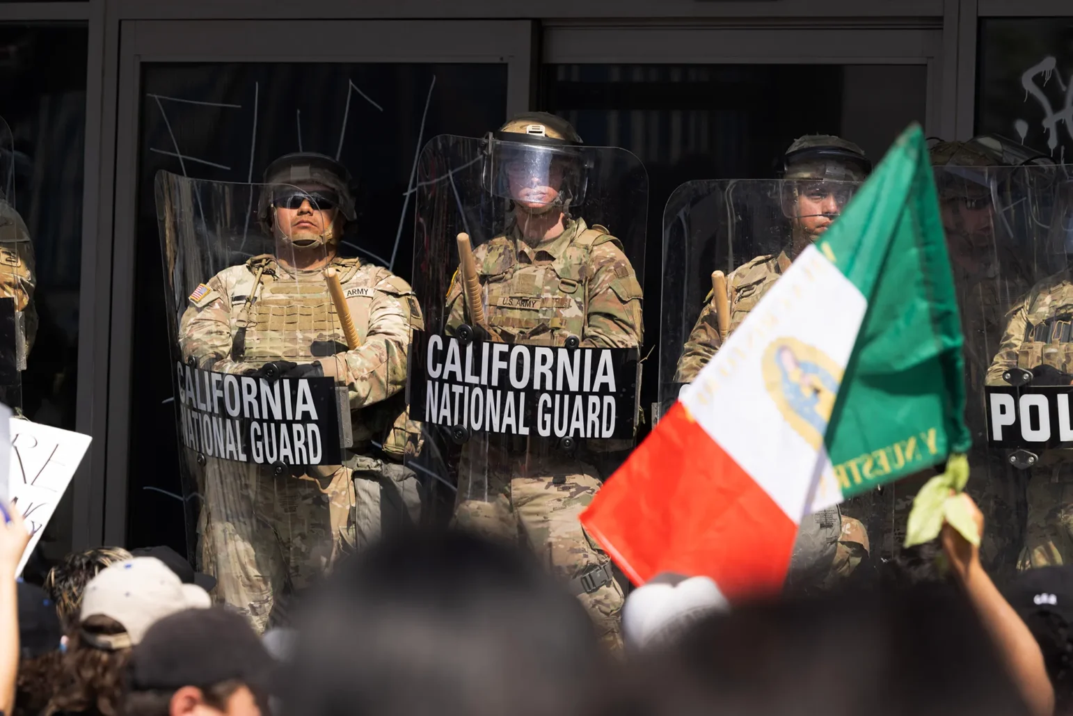 California National Guard soldiers in camouflage stand behind clear riot shields labeled “California National Guard,” holding batons as a daytime crowd protests in the foreground, with a Mexican flag and handmade signs partially visible.