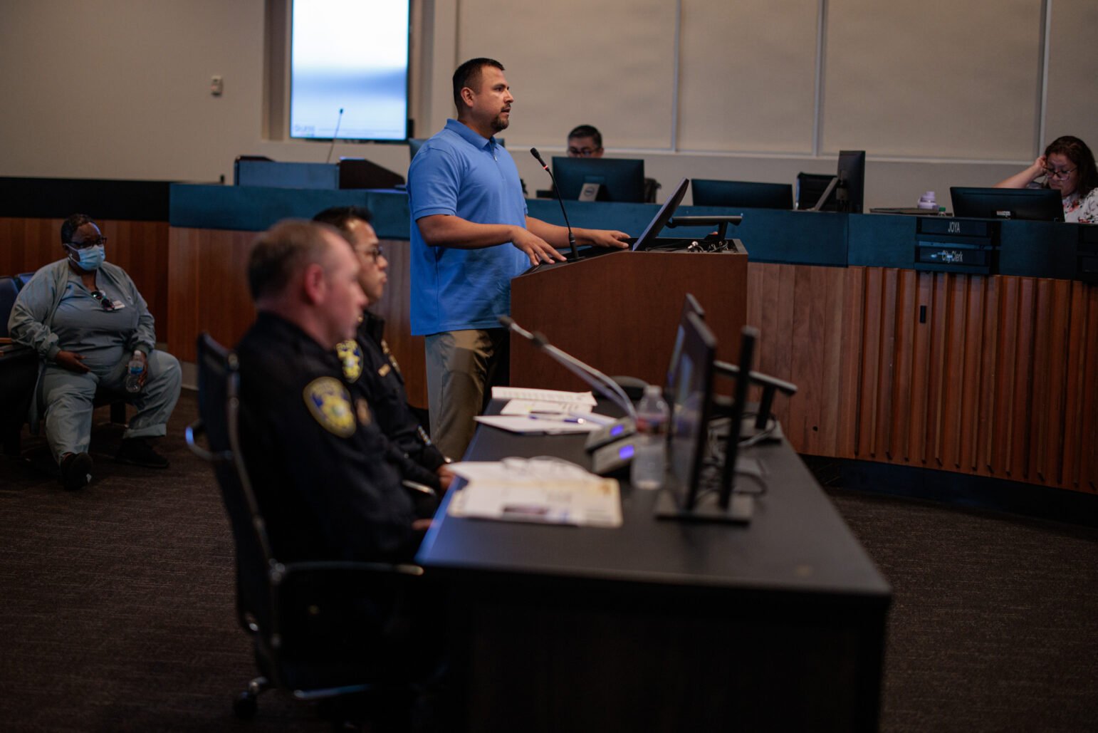 A man in a blue polo shirt speaks at a lectern during a city council meeting. Two senior police officers sit at a desk in the foreground.