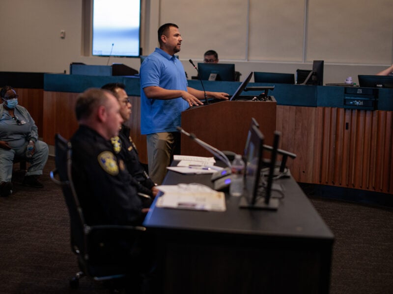 A man in a blue polo shirt speaks at a lectern during a city council meeting. Two senior police officers sit at a desk in the foreground.