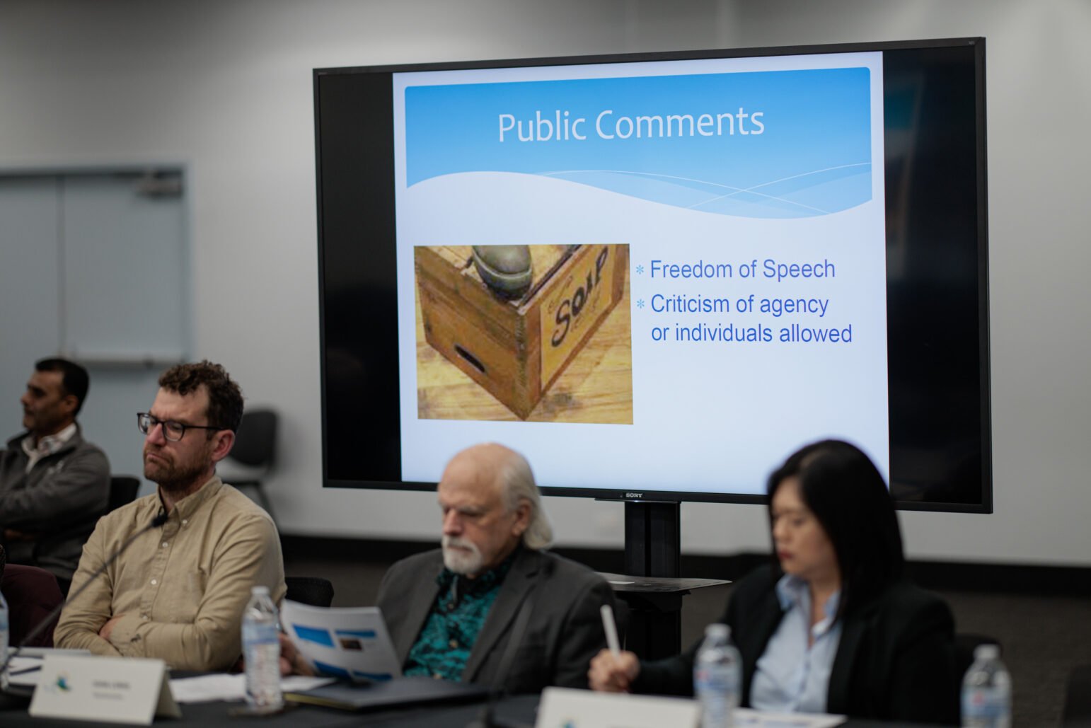 A city commission meeting with a large monitor showing a slide deck. The slide behind the commissioners reads, “Public Comments.” It features an image of a soapbox and the bullet points “Freedom of Speech” and “Criticism of agency or individuals allowed.”