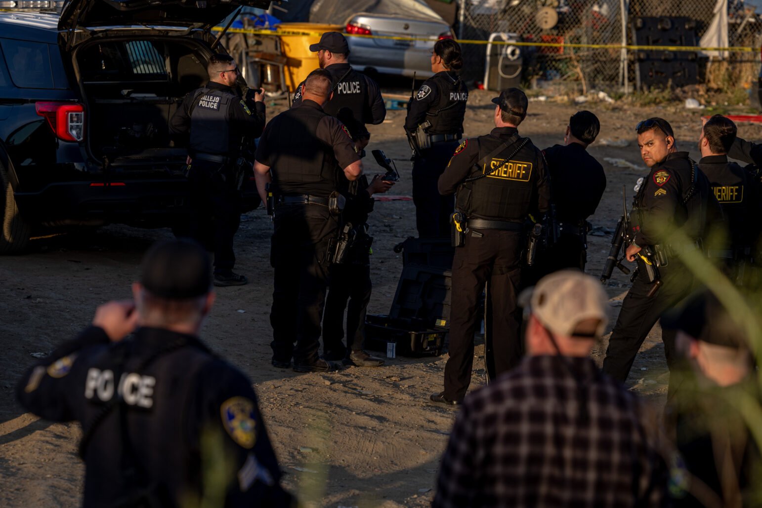 Group of police officers and sheriff’s deputies gather in a dirt lot near an open SUV, conferring, with crime-scene tape and a fenced area in the background.