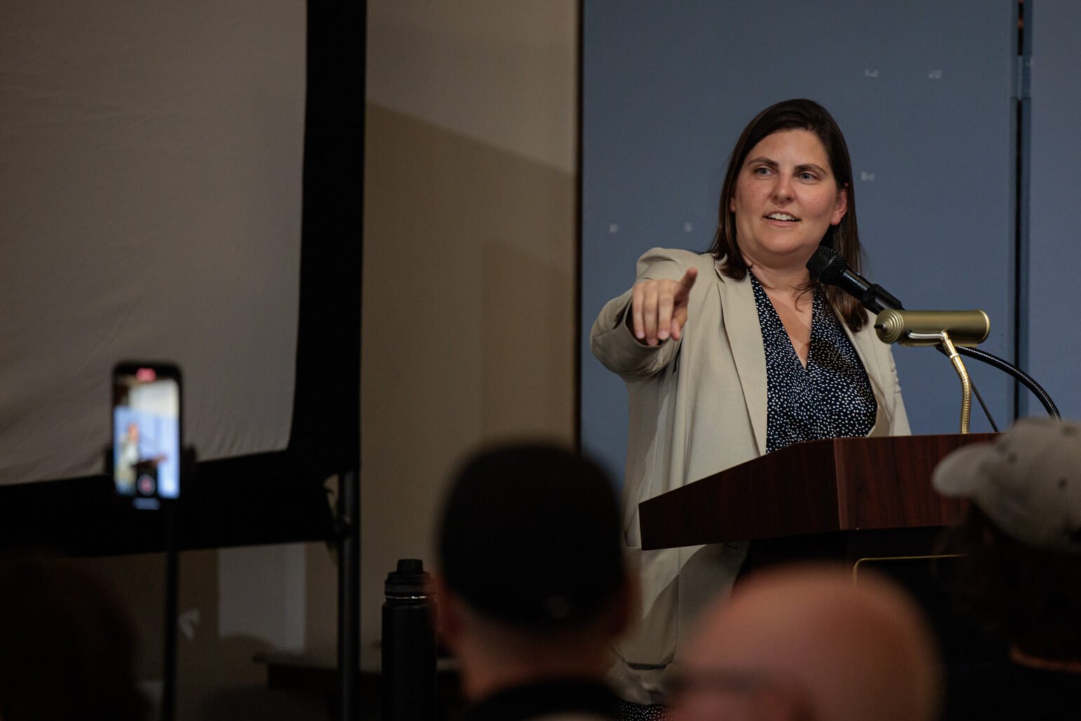 A woman in professional attire speaks at a lectern. She points toward the audience as if to take a question. A phone mounted on a tripod records her talk.