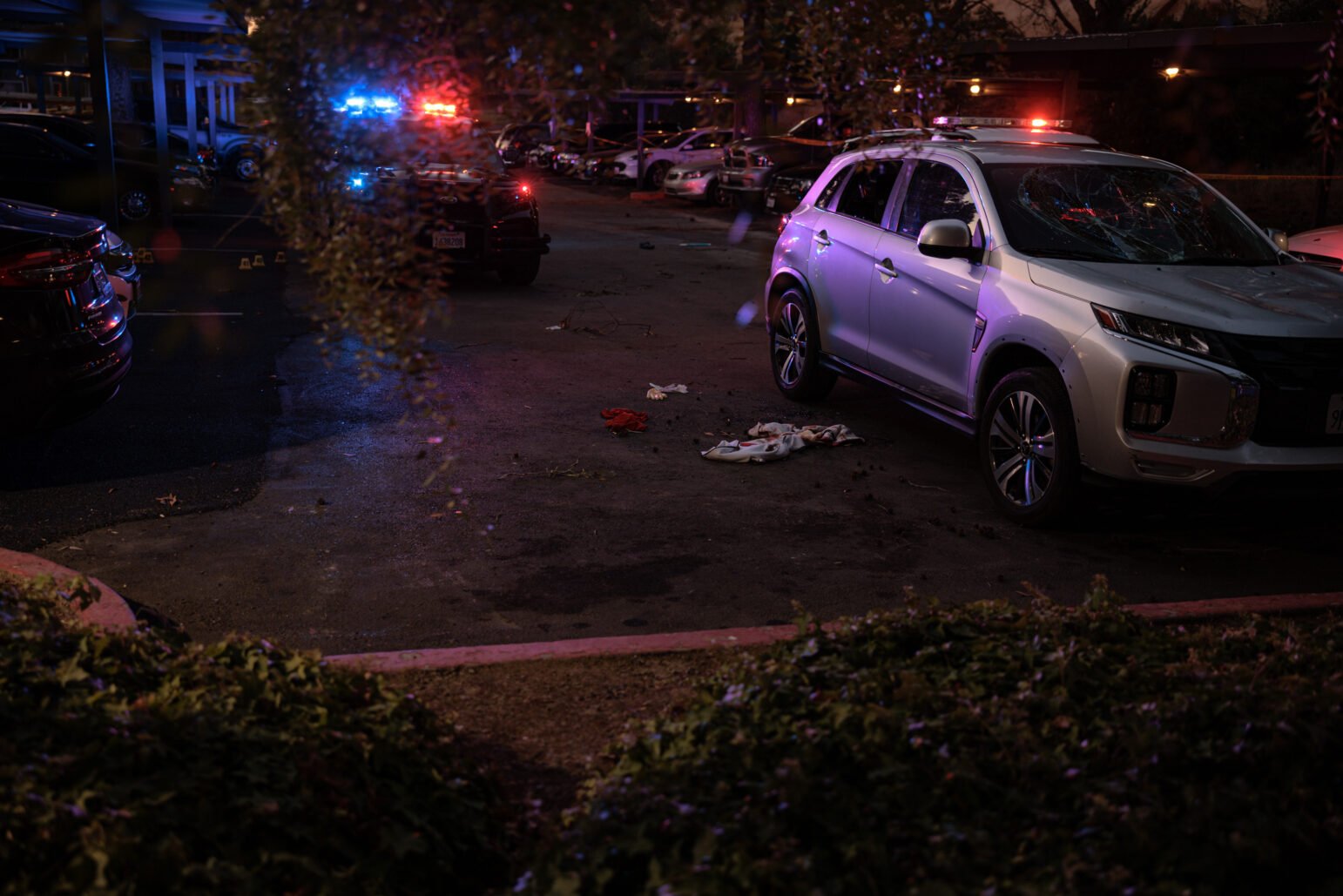 Police SUVs with flashing red and blue emergency lights illuminate a parking lot in the early evening. Clothing and other items are scattered on the ground next to a silver SUV with a shattered windshield.
