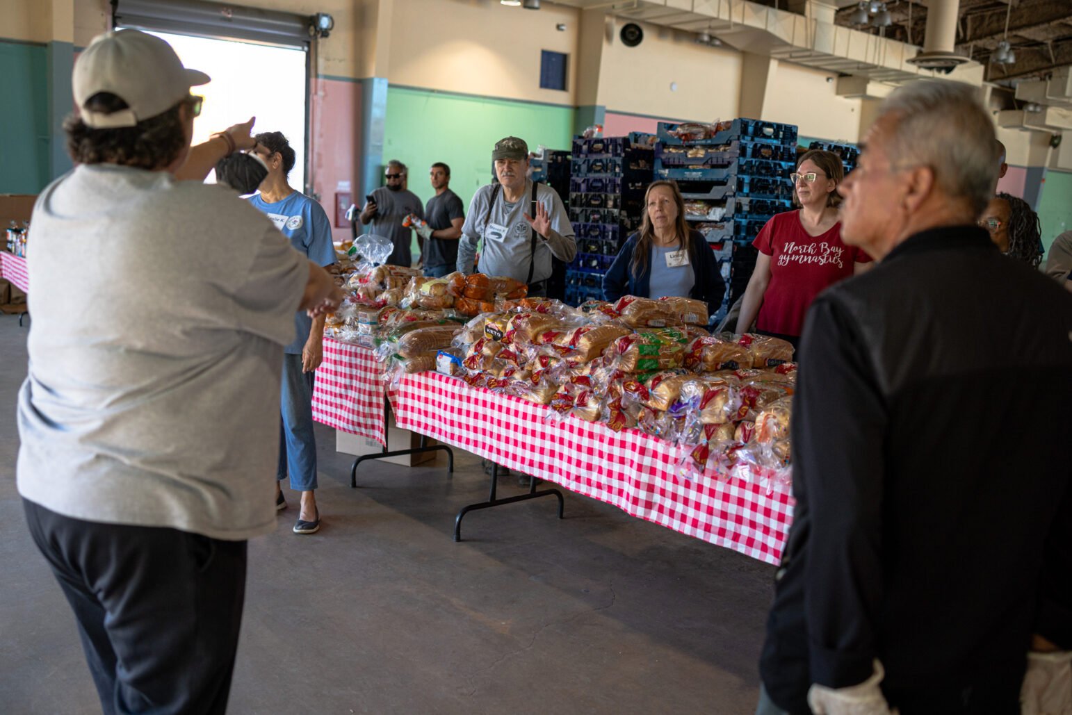Volunteers stand behind red-and-white checkered tables piled with bagged bread as a coordinator points directions at a community food distribution.