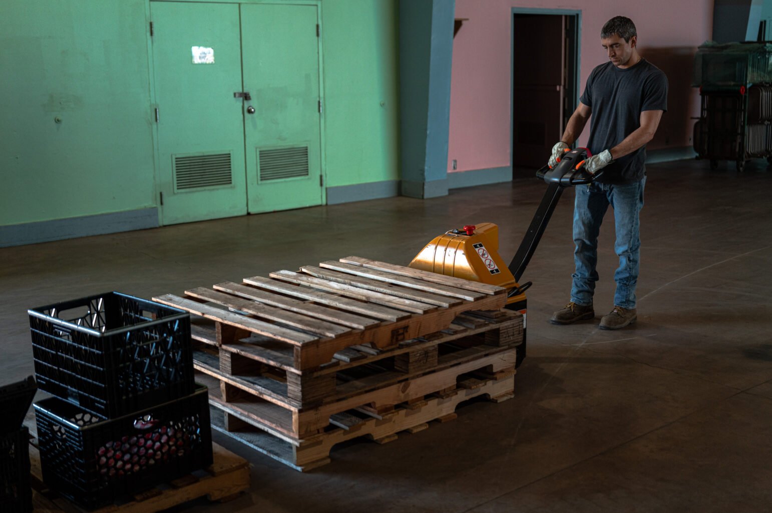 Worker in gloves uses an electric pallet jack to move stacked wooden pallets across an empty hall.