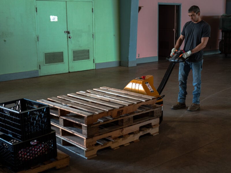 Worker in gloves uses an electric pallet jack to move stacked wooden pallets across an empty hall.