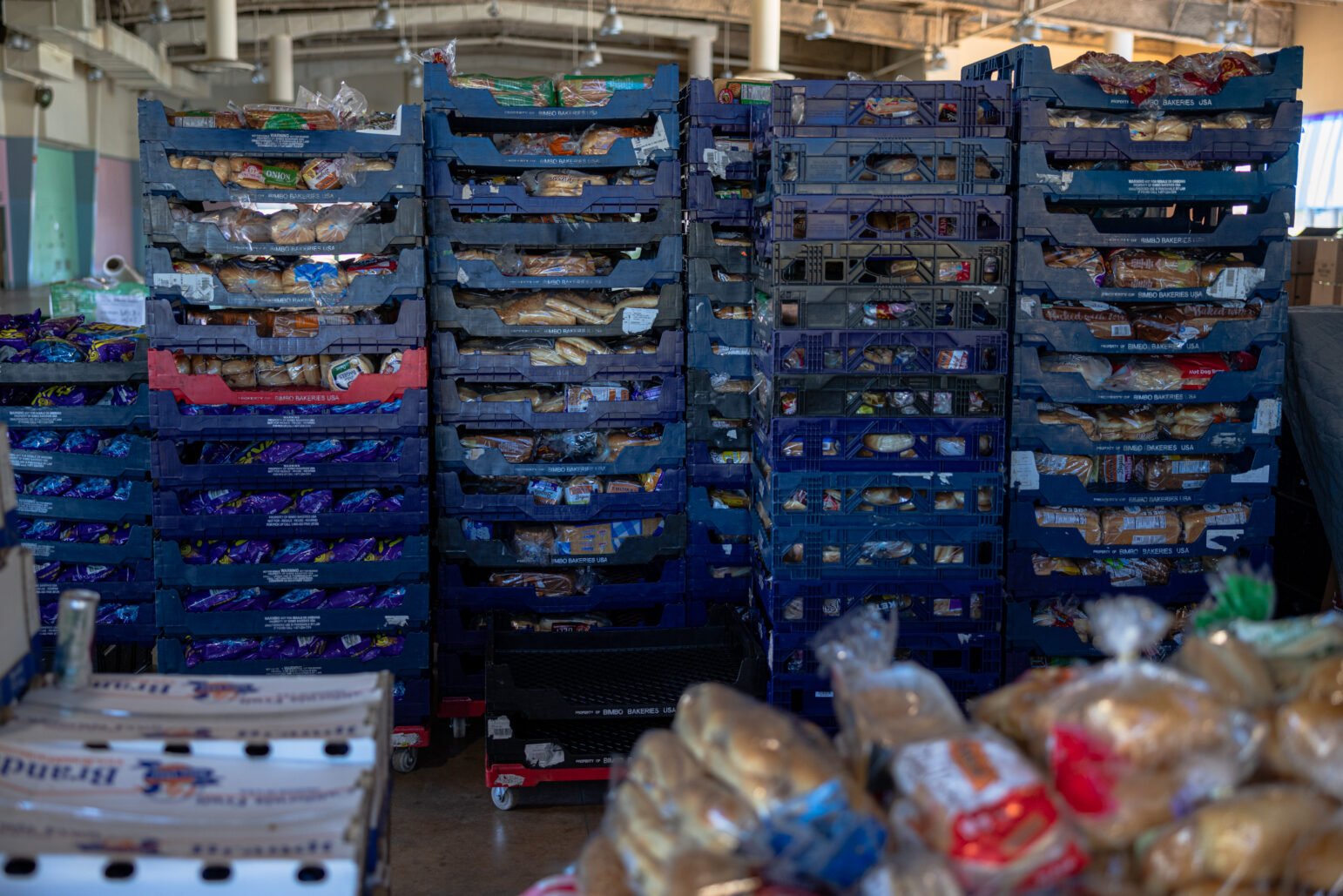 Tall stacks of blue plastic bread trays filled with packaged loaves in a storage area.