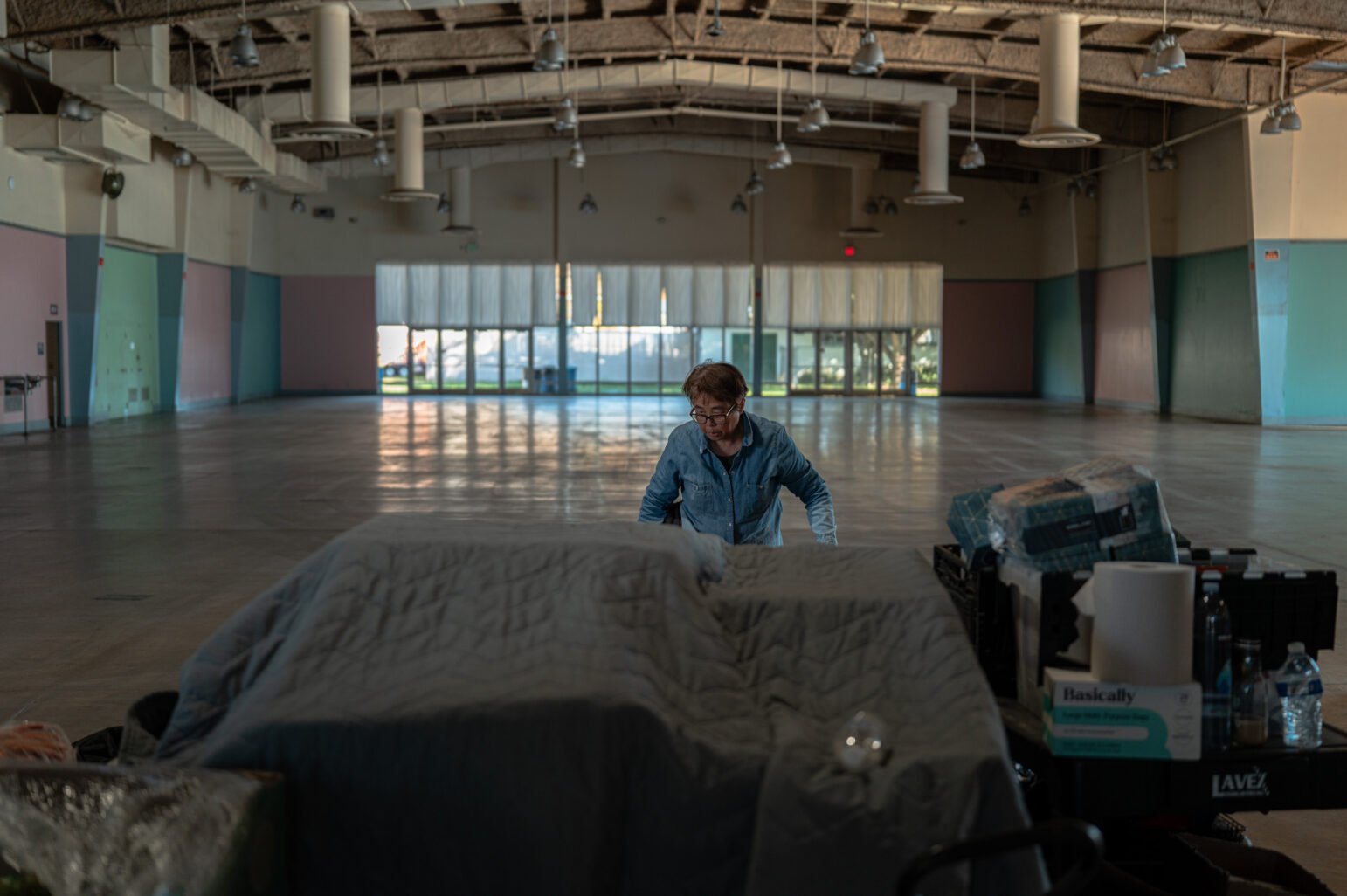 A woman organizes food supplies in a vast event hall with high ceilings and windowed doors, preparing the site.