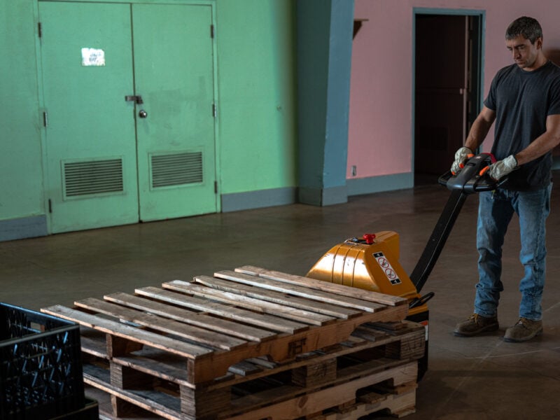 Worker in gloves uses an electric pallet jack to move stacked wooden pallets across an empty hall.