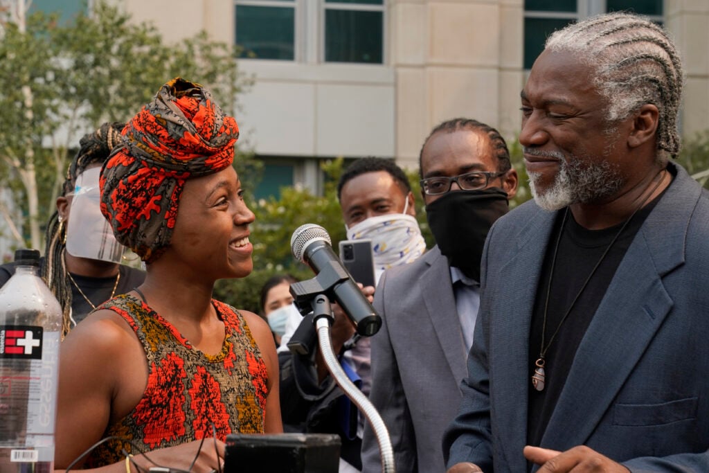 Outdoor press event: a woman in a patterned headwrap smiles beside a microphone while speaking with a man in a blazer with braided hair; masked onlookers gather behind in front of an office building.